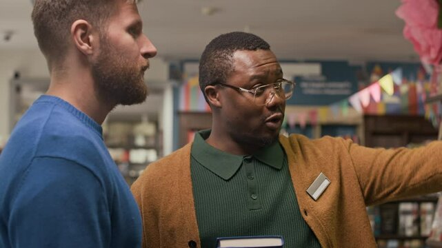 Chest up of smiling Black male bookstore worker holding book and animatedly talking to customer, standing together in colorful and welcoming shop environment