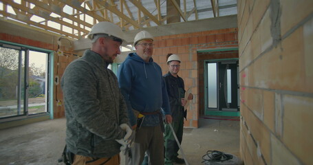 Fototapeta premium Construction workers discussing plans on site, three workers in hard hats and safety gloves reviewing blueprints in unfinished building interior