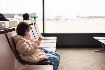 Adorable kid traveler in a winter jacket waits alone at the airport, engrossed in a mobile device. The blurred airplane view highlights solitude, digital connectivity, and modern travel habits.