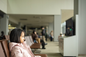 Asian adorable kid girl traveler in winter jacket sits alone in airport lounge, lost in thought. The stillness, vacant chairs, and soft lighting capture solitude, patience, and the journey ahead.