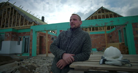 Man leaning on wooden pallets at construction site, helmet and gloves placed beside, looking into...