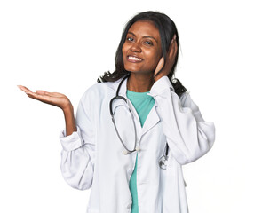 Fototapeta premium Young Indian doctor with stethoscope holds copy space on a palm, keep hand over cheek. Amazed and delighted.