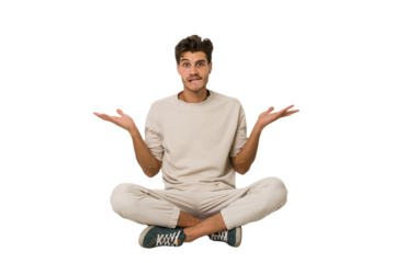 Young caucasian man sitting on the floor isolated on white background confused and doubtful shrugging shoulders to hold a copy space.