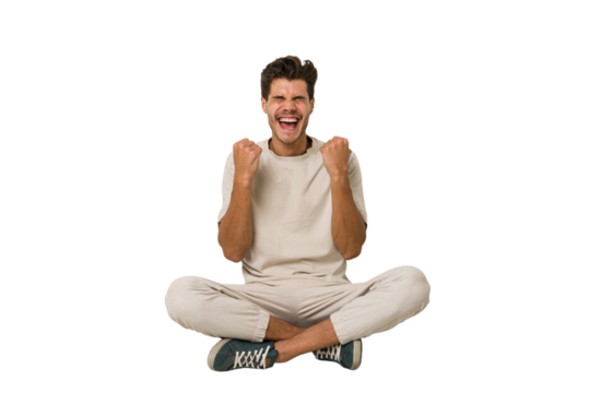 Young caucasian man sitting on the floor isolated on white background cheering carefree and excited. Victory concept.