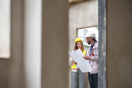 Man and Woman construction workers discussing architectural plans at a building site, standing inside a concrete structure under development reflecting teamwork
