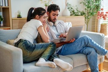 Fototapeta premium Young couple sitting on couch, engaged in laptop activity, sharing joyful moment together. cozy living room features plants and soft lighting, creating warm atmosphere