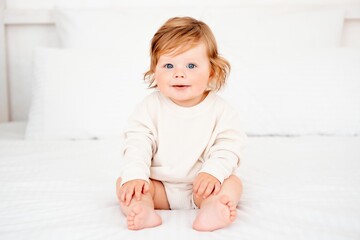 A baby with red hair and blue eyes in a white bodysuit is smiling happily on the bed at home, a...