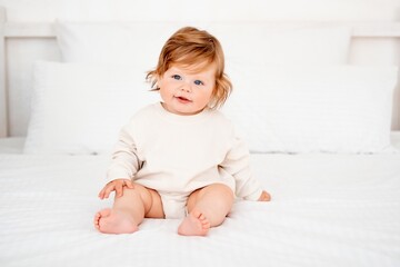 A baby with red hair and blue eyes in a white bodysuit is smiling happily on the bed at home, a...