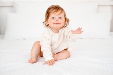 A baby with red hair and blue eyes in a white bodysuit is smiling happily on the bed at home, a...