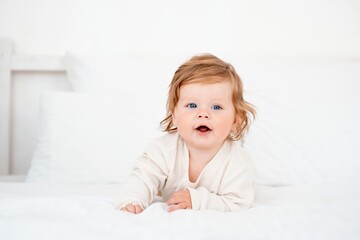 Fototapeta premium A baby with red hair and blue eyes in a white bodysuit is smiling happily on the bed at home, a healthy happy cute newborn baby boy