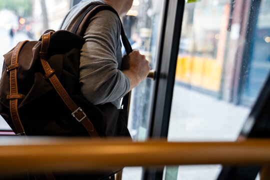 A person holding a brown backpack, getting off a bus while gripping the door handle. The image captures the typical commuter moment in an urban setting
