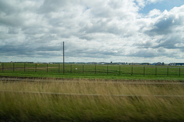 A Wide Grassy Field and Distant Airport Buildings Appear With Motion Blur Through a Tour Bus Window En Route to the Countryside Under a Cloudy Sky and Soft Natural Light.