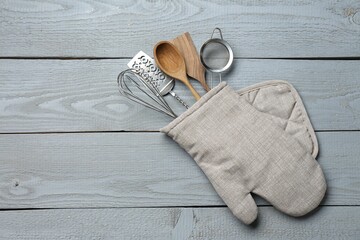 Different kitchen utensils in oven mitten on light grey wooden table, top view. Space for text