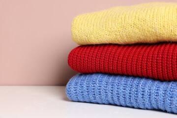 Stack of woolen sweaters on white table against color background, closeup. Space for text