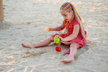 a child plays in the sandbox on the playground in summer, a little girl with bright pigtailed hair...
