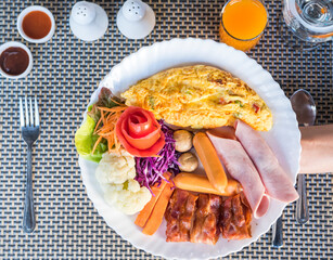 A plate with an omelet, bacon, sausage, ham, vegetables, and a side of orange juice.