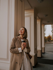 Young girl smiling and standing with coffee in her hands near a column in the city