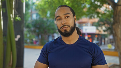 Young man with beard standing in outdoor urban park setting, wearing a blue shirt with lush green...