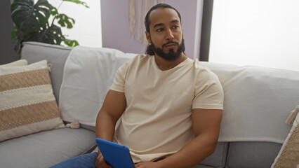 Fototapeta premium Young man with beard sitting in a cozy living room holding a tablet, surrounded by pillows and houseplants, exuding a relaxed and thoughtful atmosphere.
