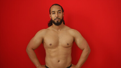 Fototapeta premium Young man with beard standing confidently against a vibrant red background highlighting his muscular physique and focused expression.