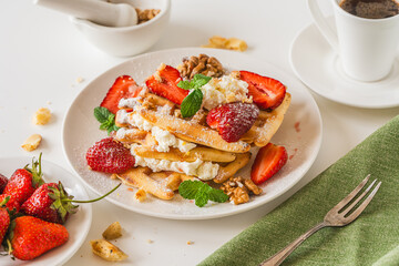 Belgian waffles with cottage cheese cream, strawberries, walnuts and mint on a white plate on a light table with a cup of coffee. Delicious, Sunday breakfast.