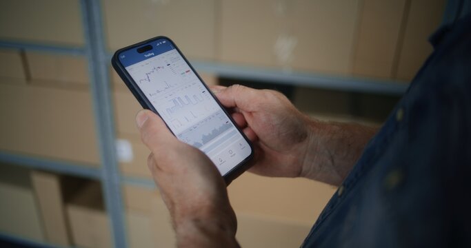 Worker Monitoring Real-Time Stocks, Exchange Market Charts Using Smartphone. Man Making Cryptocurrency Investments During Working Day in Warehouse, Combining Job with Online Trading. Close Up Shot. - Powered by Adobe