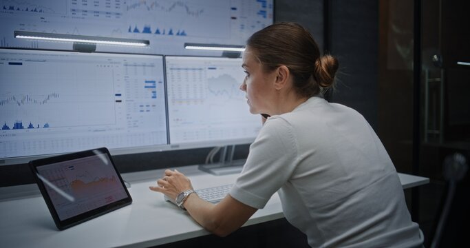 Female Financial Analyst Having Phone Call, Analyzing Real-Time Stocks, Exchange Market Charts on Multi-Monitor Computer and Digital Tablet. Businesswoman Working in Financial Agency Office at Night.