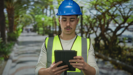 Young hispanic man wearing hardhat and reflective vest uses tablet in a lush outdoor park environment, embodying modern construction and technology synergy.