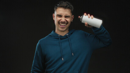 Young man holding spray paint can against isolated black background, wearing blue hoodie,...