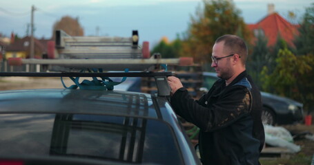 Worker securing ladder onto car roof rack, preparing for transportation, outdoor setting,...