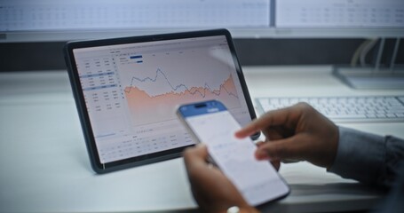 African American Stock Exchange Broker Analyzing Real-Time Stocks and Exchange Market Charts on Smartphone and Digital Tablet Using Online Trading App. Investment Agent Working in Office. Close Up.
