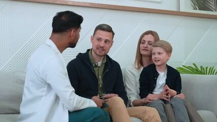 Indian doctor showing something on a tablet to young parents with their son, sitting on a sofa in a hospital waiting room, talking about diagnosis and treatment