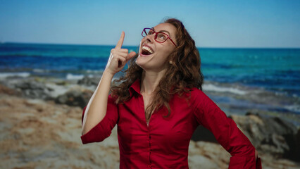 Woman with red shirt joyfully laughing by seaside, pointing upwards against clear blue sea...