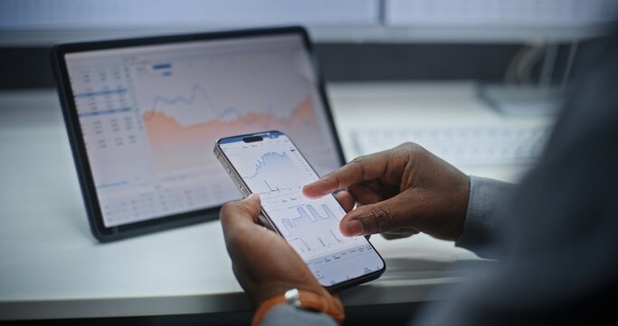 African American Stock Exchange Broker Analyzing Real-Time Stocks and Exchange Market Charts on Smartphone and Digital Tablet Using Online Trading App. Investment Agent Working in Office. Close Up.