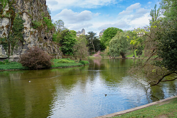 Scenic spring view of lake, cliffs, and Haussmann buildings in Parc des Buttes-Chaumont, Paris, France. Urban nature and relaxing park landscape with water and greenery.