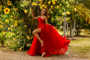 Elegant woman in flowing red dress poses gracefully among yellow tropical flowers and lush greenery...