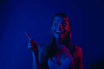 Portrait of a happy young woman in neon lighting pointing excitedly against a vibrant blue and pink...