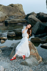 Serene woman in white dress and red shoes sitting on rocky shoreline by the ocean