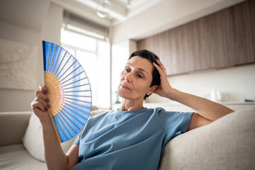 Exhausted mature woman fanning herself at home due to unbearable heat. Elderly suffering from...