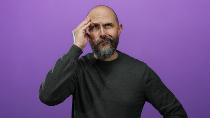 Obraz premium Bald man with a beard in a gray sweater looks puzzled with a hand on his forehead against an isolated purple background.