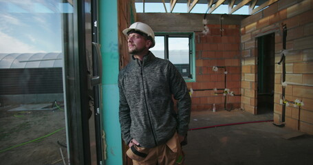 Young construction worker gazing outside, hands in pockets, hard hat on, inside partially constructed building, with unfinished brick walls and window frame in background