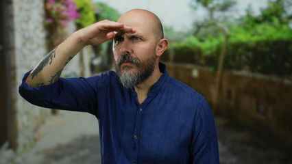 Fototapeta premium Man with beard and tattoo looking into the distance on a city street wearing a blue shirt on a sunny day outside with greenery and flowers