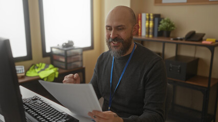 Man with beard reviewing documents at desk in modern office featuring computer monitor and shelves in background with books and decor creating a professional workspace atmosphere.