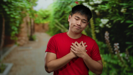Fototapeta premium Young man in red shirt holding chest in park setting with green lush background, displaying emotion and introspection outdoor.