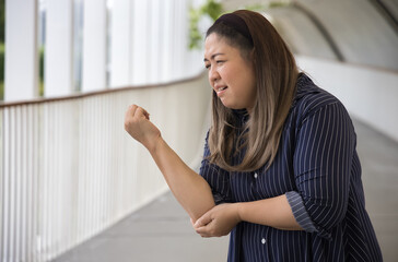 Plus-sized chubby Asian middle-aged aunt woman suffering from tennis elbow syndrome or elbow joint pain or injury, injured elbow tendon, rheumatoid or gout arthritis concept