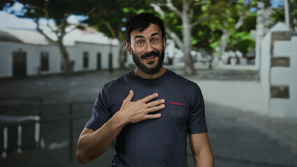 Fototapeta premium Man with beard standing outdoors on a sunny street background expressing surprise with hand on chest