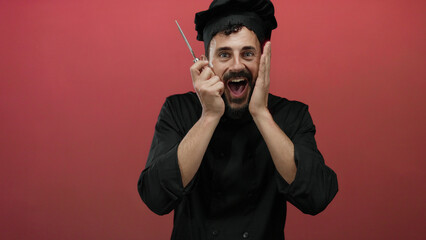 Man with beard wearing chef outfit enthusiastically holding cheese grater, standing before red...