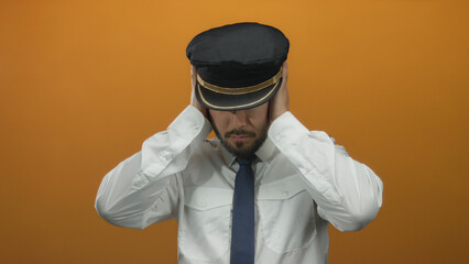 Young man in pilot uniform with hat covers his ears against an orange background, emphasizing a sense of frustration or distress.