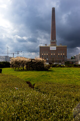 30.03.2025 Gent, Belgium, a tall brick smokestack rises from a Luminus power station under dramatic cloudy skies with green lawns, trimmed bushes, and blooming white trees in the foreground.
