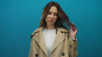 Woman wearing beige coat over isolated blue background playing with her hair, expressing curiosity...